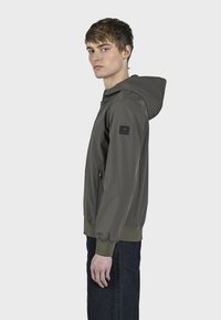 Maslinasto zelena dukserica sa zatvaračem sprijeda, sa logotipom na rukavu, rebrastim manžetama i jednostavnim, uskim dizajnom.