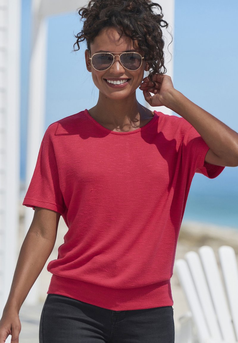 Rotes T-Shirt mit kurzen Ärmeln, lässigem Schnitt und rundem Ausschnitt, kombiniert mit dunklen Jeans. Das Material wirkt leicht und strukturiert.