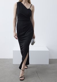 Robe noire à épaule unique avec un corsage ajusté et une jupe drapée avec une fente sur le côté. Le mannequin tient une pochette métallique et porte des sandales noires.