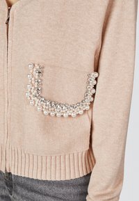 Strickjacke in hellpeach mit einem Reißverschluss, ausgestattet mit einer durch Perlen und funkelnden Details verzierten gepunkteten Tasche. Rippbündchen.