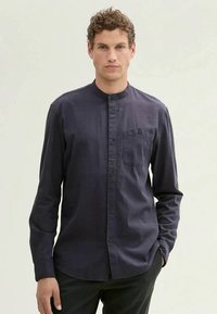 Uomo con capelli ricci castani che indossa una camicia a colletto mandarino blu scuro con bottoni e una tasca sul petto, in piedi di fronte a uno sfondo chiaro e semplice.