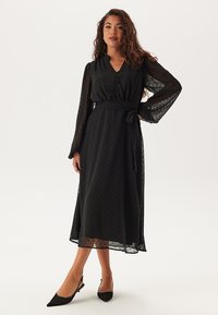 Svart midiklänning med prickigt mönster, långa genomskinliga ärmar, v-ringning, främre knappar och midjeband. Mjuk, lätt tyg med flödande design.