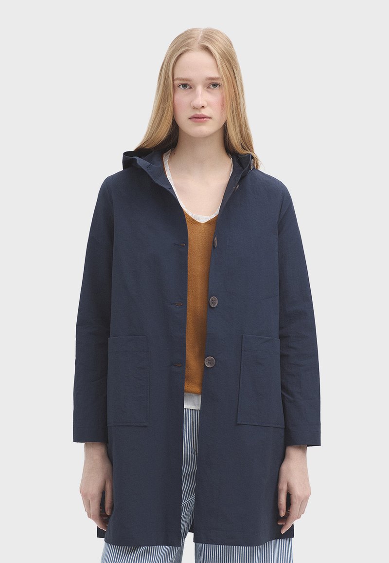 Námořnický bavlněný kabát s kapucí, předními knoflíky, dvěma velkými kapsami a rovný střih, doplněný hnědým topem a pruhovanými kalhotami.