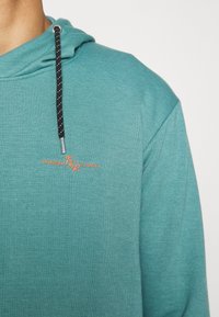 Felpa color teal con tessuto testurizzato, coulisse con un motivo bianco e nero e un logo ricamato arancione sul davanti.