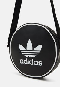 Borsa rotonda nera in pelle con dettagli bianchi, che presenta un logo Adidas bianco di grandi dimensioni e un design a tre strisce. Spallaccio nero regolabile.