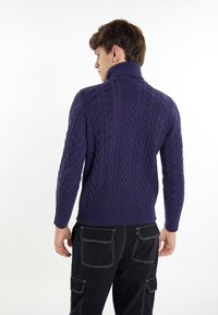 Maglione viola a treccia con un colletto alto e orlo a coste. Texture dettagliata con un motivo di treccia molto evidente. Indossato con pantaloni scuri.