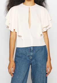 Różowa bluzka z falbaniastego rękawa, dekoltem w kształcie łezki i skróconym fasonem, zestawiona z dżinsami z wysokim stanem. Gładka struktura i lekki materiał.