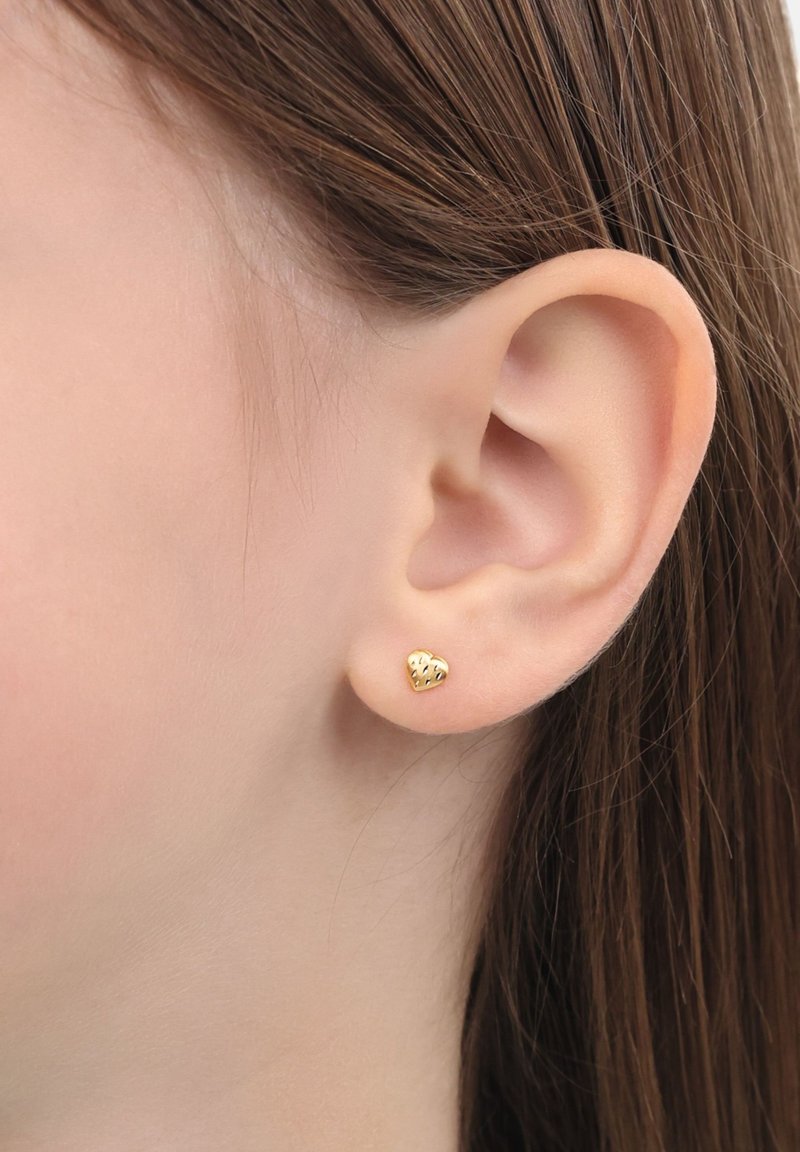 Pendiente de tuerca en forma de corazón dorado con superficie texturizada, mostrado en la oreja de una persona con cabello castaño liso. Diseño simple y compacto.