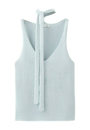 Młoda kobieta z długimi włosami ubrana w jasnoniebieski, dziergany, bezrękawnik, szerokie niebieskie dżinsy i czarne klapki, stojąca z rękami w kieszeniach.