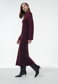 Vestido burdeos de manga larga con rayas negras horizontales, silueta ajustada y dobladillo acampanado. Combinado con botas negras de punta alta hasta la rodilla.