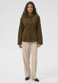 Woman with long wavy hair wearing an olive buttoned corduroy shirt, beige straight-leg pants, and black pointed shoes, standing against white background.