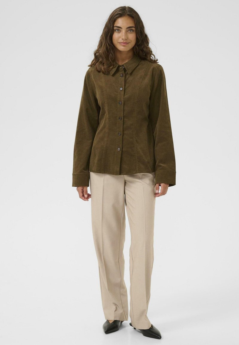 Woman with long wavy hair wearing an olive buttoned corduroy shirt, beige straight-leg pants, and black pointed shoes, standing against white background.