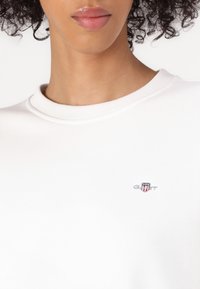 Weißes Sweatshirt mit Rundhalsausschnitt aus glattem Stoff. Verfügt über ein kleines, gesticktes Logo auf der Brust. Dezente gerippte Struktur am Kragen.