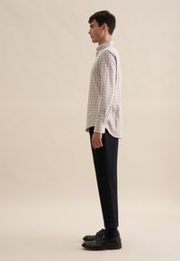 Camicia a maniche lunghe a quadri da uomo in bianco, marrone e beige; indossata con pantaloni scuri e scarpe nere, evidenziando una vestibilità rilassata e i polsini.