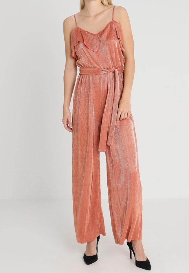 Mono de color rosa hecho de una tela texturizada y brillante. Presenta un escote con volantes, cintura ceñida con cinturón y un diseño de pierna ancha. Se llevan tacones negros.
