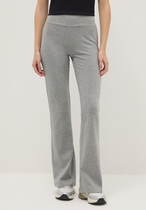 Femme portant un pantalon gris clair à jambes droites et des baskets argentées, debout devant un fond beige uni.
