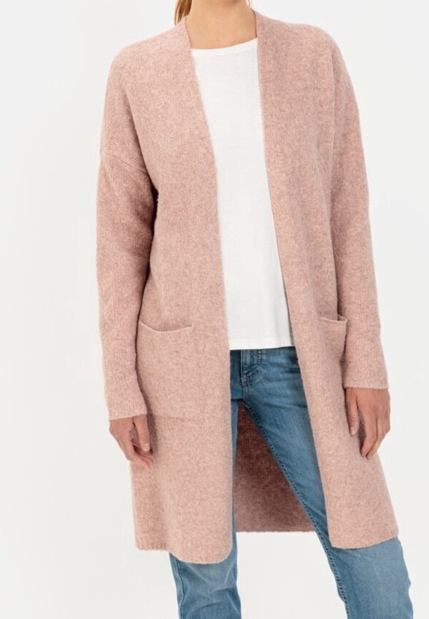 Femme portant un long cardigan rose poudré ouvert devant avec des poches avant, associé à une chemise blanche et un jean bleu.