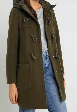 Femme portant un manteau vert olive à brandebourgs avec de grandes poches avant, une capuche, un pull gris et un jean bleu, debout devant un fond neutre.