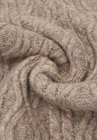 Tessuto a maglia cablè in una morbida lana beige testurizzata, che mostra un motivo attorcigliato con fibre visibili e una caduta delicata.
