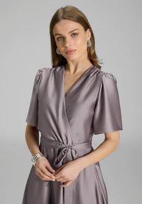 Lavendel satijnen wikkeljurk met korte pofmouwen en een ceintuur in de taille. Heeft een V-halslijn en een vloeiende rok. Zilveren armbanden en oorbellen.