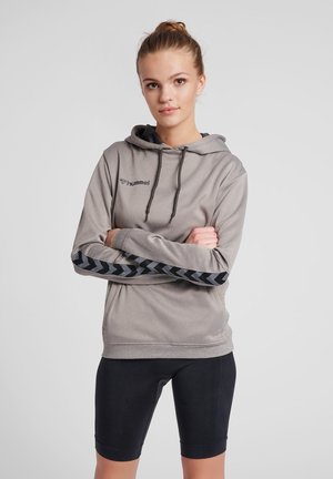 Sweatshirt à capuche gris avec texture côtelée, présentant des accents en chevrons noirs sur les manches. Associé à un short de cyclisme noir. Logo sur la poitrine.