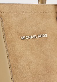 Tako hnědá semišová taška s kovovým logem "MICHAEL KORS", hladkým povrchem a kontrastním prošíváním kolem okrajů.