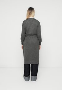 Langer, grauer, strukturierter Cardigan mit geraffter Taille. Verfügt über gerippte Bündchen und Saum, kombiniert mit dunkelblauen hochgekrempelten Jeans.