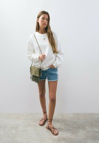 White sweatshirt with a star graphic, denim shorts with frayed hems, olive green fringed bag, and brown strappy sandals. Neutral background.