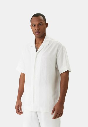 Uomo di colore che indossa una camicia bianca a maniche corte con bottoni e pantaloni bianchi, in piedi davanti a uno sfondo chiaro e semplice.