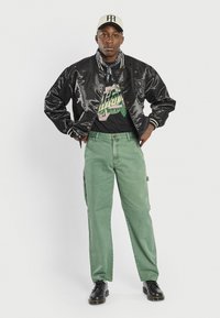 Jachetă bomber din satin negru cu manșete în dungi, tricou grafic negru, pantaloni cargo verzi și pantofi negri. Inclusiv o șapcă neutră și un eșarfă patterned.