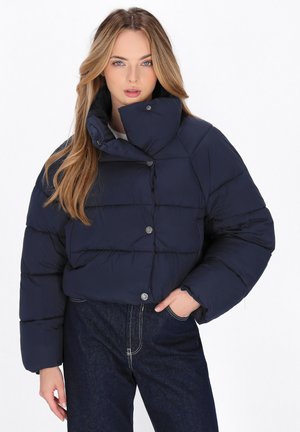 Marineblauwe pufferjack met hoge kraag, knoopsluitingen en een gestikt ontwerp, gecombineerd met donkerblauwe jeans. Glad materiaal, oversized pasvorm.