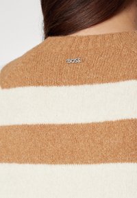 Jasnobeżowy sweter w brązowe paski, wykonany z teksturowanej dzianiny, z prążkowanym dekoltem i srebrnym logo "BOSS" na plecach.