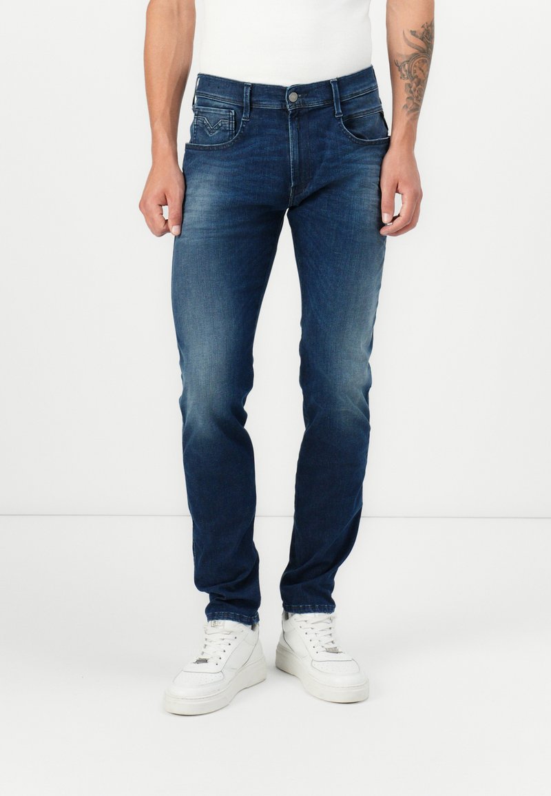 Tummansiniset slim fit -farkut, joissa on viisi taskua ja minimaalinen kulutus. Yhdistetty valkoisiin korkeisiin tennareihin.