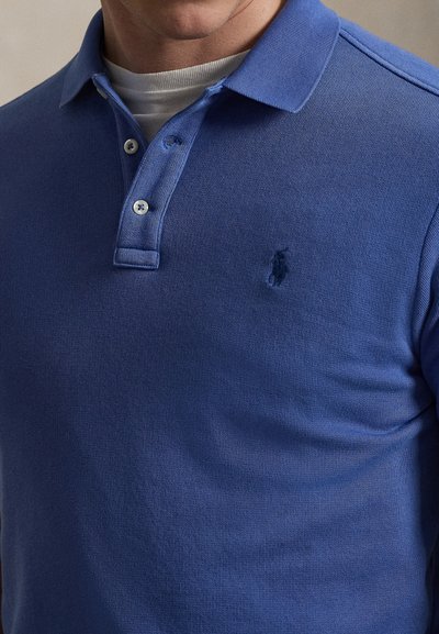 Άνδρας που φοράει μπλε πόλο μπλουζάκι με λευκό εσώρουχο, με γιακά που κουμπώνει και μικρό κεντημένο λογότυπο στο στήθος.