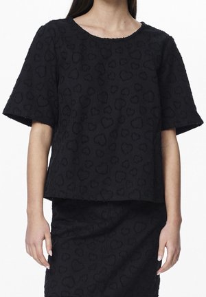 Donna che indossa un top corto a maniche corte nero dalla texture e una gonna abbinata con un sottile motivo a cuori su uno sfondo bianco uniforme.