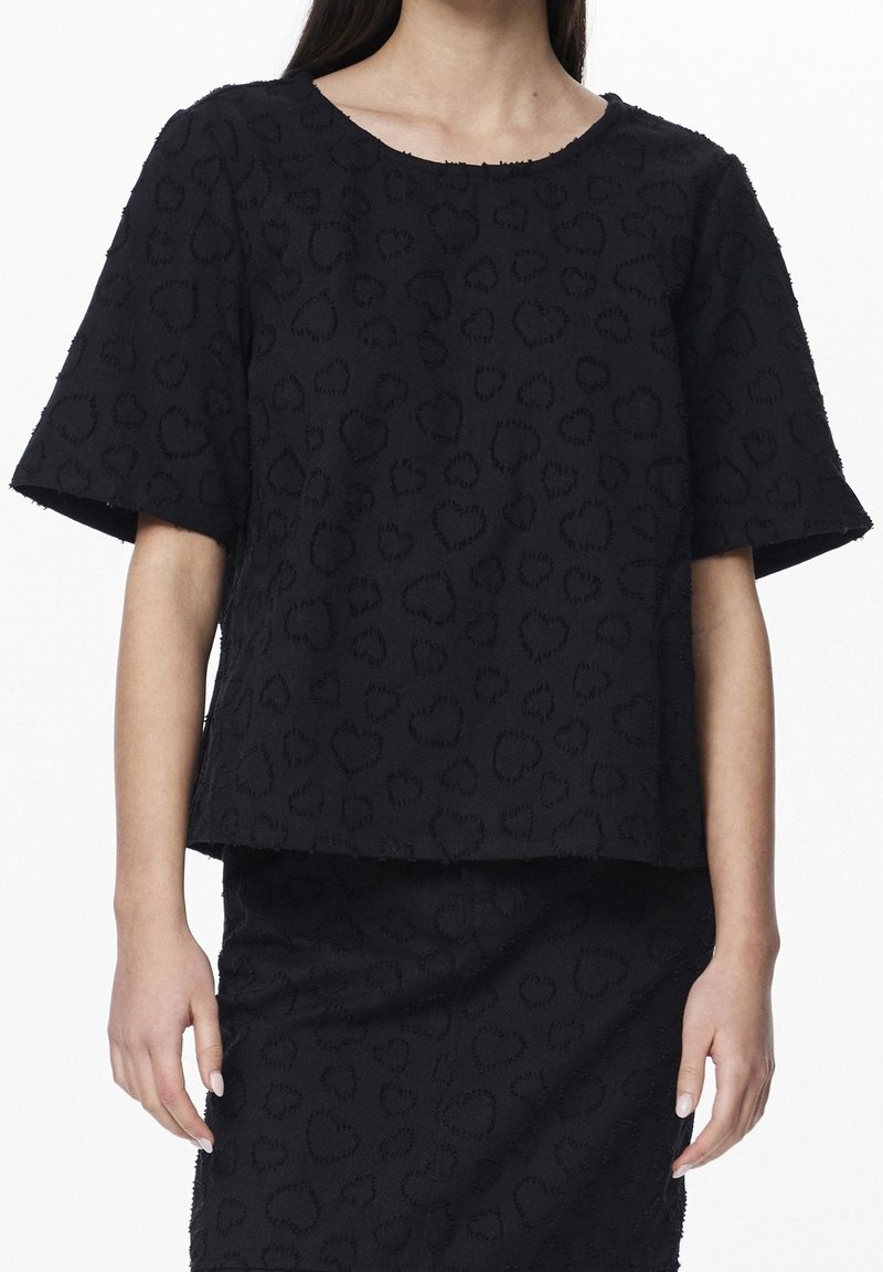 Woman wearing a black textured short-sleeve top and matching skirt with a subtle heart pattern on a plain white background.