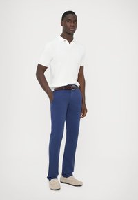 Kortermet hvit polo med teksturert mønster, kombinert med blå bukser og et brunt flettet belte. Beige slip-on sko fullfører antrekket.