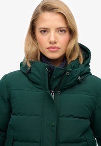 Zielona pikowana kurtka z wysokim kołnierzem i regulowanym kapturem. Posiada przedni zamek błyskawiczny, guziki oraz teksturowaną dzianinową podszewkę wewnętrzną.