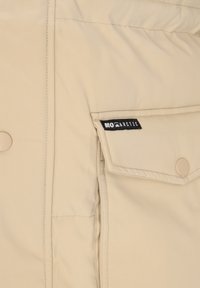 Beżowa ocieplana kurtka z boczną kieszenią z czarną metką, gładką fakturą, zapięciem na guziki i subtelnymi detalami szwów.