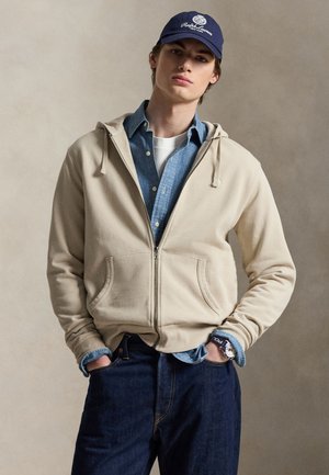 Noor mees kannab beeži lukuga dressipluusi sinise nupuga särgi ja valge T-särgi peal, tumedad teksad, tumesinist Ralph Laureni mütsi ja käekell tumesinise rihmaga.