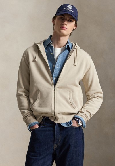Jeune homme portant un sweat à capuche zippé beige sur une chemise bleue à boutons et un t-shirt blanc, jean foncé, casquette Ralph Lauren bleu marine et montre-bracelet avec un bracelet bleu marine.