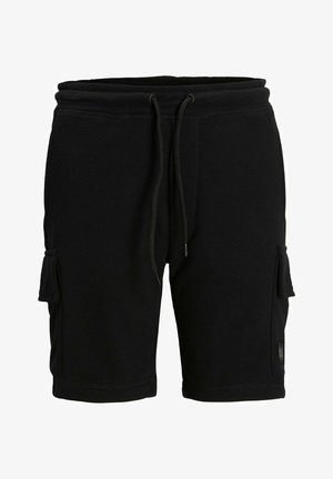 Dziecko stojące, ubrane w białą koszulkę z napisem "JACK & JONES", czarne szorty, czarne skarpetki i białe sneakersy, na jednolitym jasnym tle.