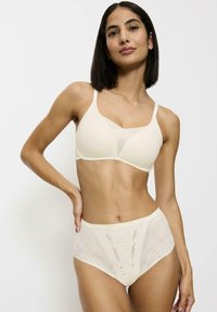 Sostén inalámbrico beige con una textura suave y un diseño que realza la figura. Las braguitas altas y transparentes presentan sutiles patrones ondulados. Ambos comparten colores similares.