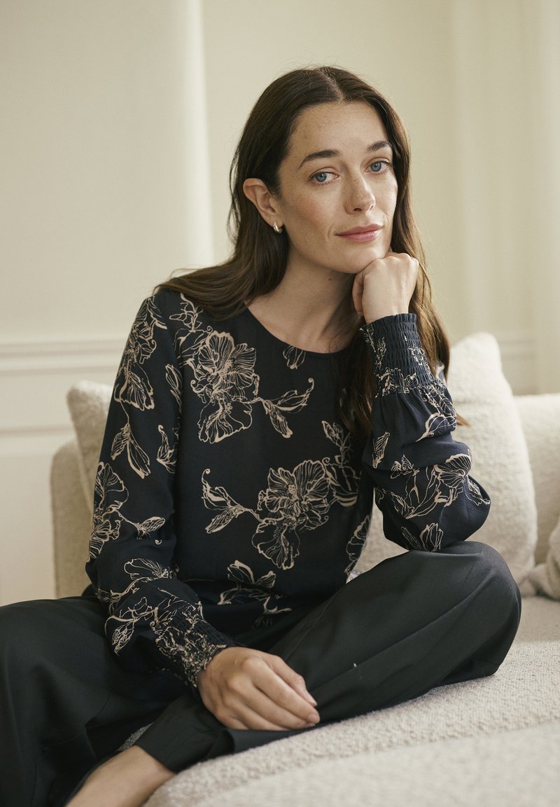 Hosszú, barna hajú nő keresztbe tett lábakkal ül egy kanapén, állát a kezére támasztva, fekete virágmintás blúzt és fekete nadrágot visel.