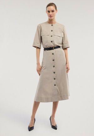 Frau trägt ein beigefarbenes Midi-Hemdkleid mit Ellbogenlangenärmeln, Brusttaschen, schwarzem Gürtel und schwarzen spitzen Pumps, stehend.