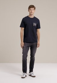 Mornarica pamučna majica kratkih rukava sa grafičkim printom na lijevom prsnom dijelu. U kombinaciji sa tamno sivim trapericama i bijelim tenisicama.