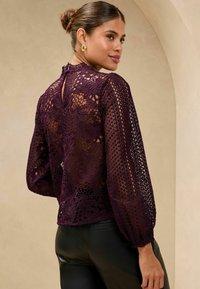 Violetti pitsilä blouse pitkillä, puffettihihoilla, kukkakuviolla ja avotakakiinnityksellä. Yhdistetty mustiin, istuviin housuihin.