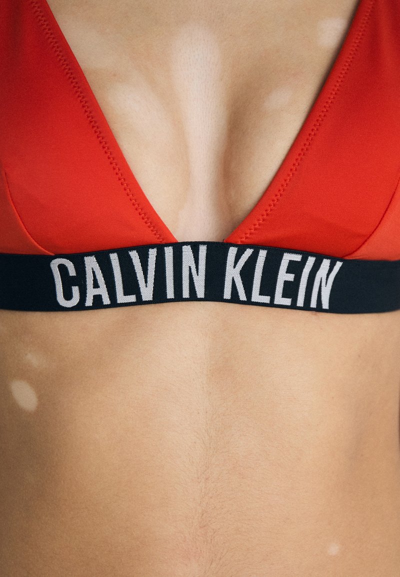 Rode bralette met een diepe V-halsontwerp en een zwarte elastische band met het "CALVIN KLEIN" logo in het wit. Soepele stofstructuur.