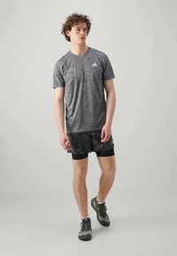 T-shirt de sport gris à col rond et manches courtes, associé à un short noir à motifs. Des chaussures de sport décontractées complètent la tenue.