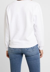 Persona che indossa un semplice maglione bianco a maniche lunghe e jeans di denim blu, fotografata di spalle su uno sfondo bianco.